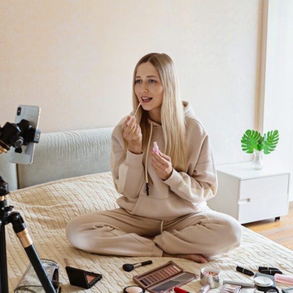 Young Caucasian beauty blogger applying makeup with decorative cosmetics on bed